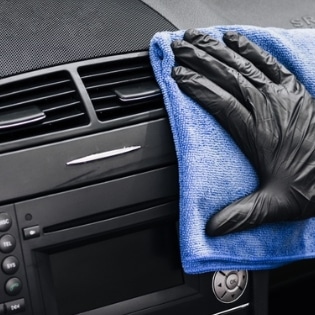 Main gantée en latex essuyant méticuleusement le tableau de bord d'une voiture avec un chiffon bleu violet, représentant le service de nettoyage de véhicule d'ISL à Mâcon